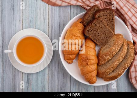 vista dall'alto di pane come croissant segale affettata e pannoccio marrone seminato nel piatto su tela e tazza di toddy caldo su sfondo di legno Foto Stock