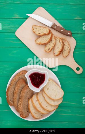 vista dall'alto di pane come croissant a fette con coltello sul tagliere e pannocchetti marroni e baguette a fette con marmellata di lamponi su sfondo verde Foto Stock