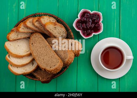 vista dall'alto di pane come pannoccio bruno seminato e fette di baguette nel cestino e marmellata di lamponi in ciotola con tazza di tè su sfondo verde Foto Stock