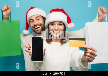 donna in santa cappello mostrando carta di credito e smartphone vicino uomo sfocato con borse shopping isolato su blu Foto Stock