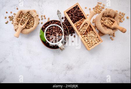 Fagioli di caffè verdi e marroni, tostati e scuri, in sacchi di canapa con sessole su fondo di cemento bianco. Foto Stock