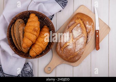 vista dall'alto di pane come croissant e fette di pane di panino bruno seminato nel cestino su un panno e pane crostoso sul tagliere su sfondo di legno Foto Stock