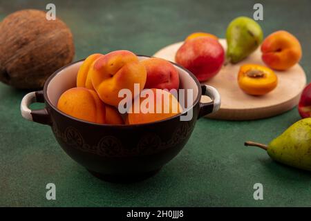 vista laterale di albicocche intere in ciotola e mezza tagliata una con pera di pesca sul tagliere e cocco su sfondo verde Foto Stock