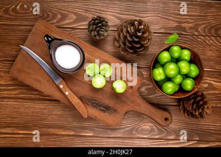 vista dall'alto delle prugne verdi affettate con sale e coltello da cucina su un tagliere in legno, coni e prugne in una ciotola su sfondo rustico in legno Foto Stock