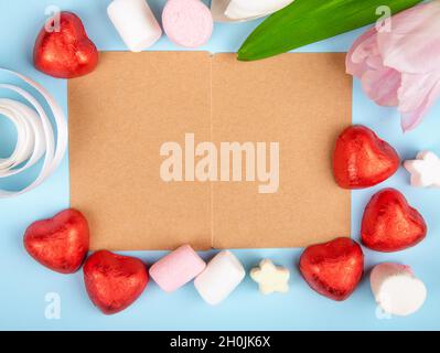 vista dall'alto di un biglietto d'auguri di carta marrone con marshmallow sparsi e caramelle al cioccolato a forma di cuore in lamina rossa con tulipani rosa su blu Foto Stock