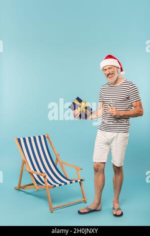 felice uomo maturo in santa hat che tiene un bicchiere di champagne e una scatola regalo vicino sdraio su blu Foto Stock