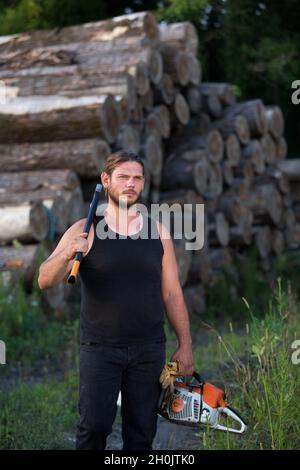 Giovane e forte lumberjack che porta l'ascia sulla spalla e motosega in altra mano di fronte a tronchi di legno su palo in foresta Foto Stock