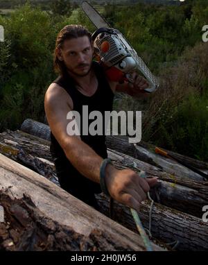 Giovane uomo forte lumberjack portare motosega a spalla e salire su un mucchio di tronchi Foto Stock