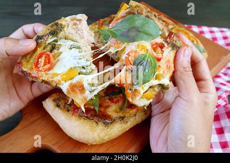 Fettine di appetitosa pizza pesto vegetariana con formaggio fuso allungato in mano Foto Stock