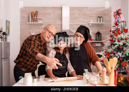 Nonno e bambino che assumono selfie il giorno di natale indossando zafferano e bonette. Felice allegra joyfull adolescente ragazza aiutare donna anziana preparare dolci biscotti per celebrare le vacanze invernali. Foto Stock