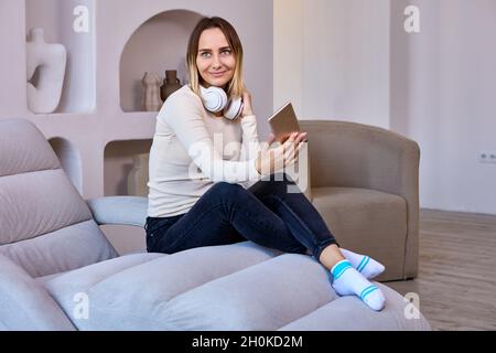 Donna felice in cuffie senza fili con telefono si siede sul divano interno. Foto Stock