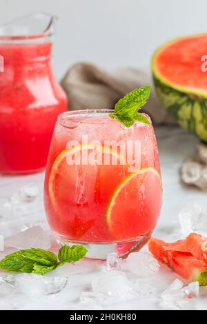 Cocomero mojito fresco bevanda rossa. Con cocomero, limone, lime, foglie di menta e ghiaccio. La bevanda è su un tavolo bianco con un mezzo cocomero sul retro Foto Stock