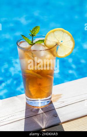 Un bicchiere di tè al ghiaccio con limone e menta, a bordo piscina con acqua blu sullo sfondo. Immagine verticale. Sul bordo del bicchiere a è presente un limone a fette Foto Stock