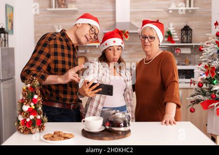 Famiglia felice che prende selfie usando il telefono che fa le espressioni divertenti durante il tempo della foto che celebra la festa di natale insieme in cucina culinaria decorata di Natale. Nipote godersi la stagione invernale Foto Stock