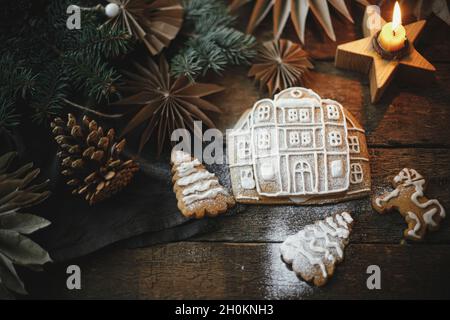 Biscotti di Natale di pan di zenzero con glassa su tavola rustica di legno con candela e ornamenti, disposizione piatta. Casa di pan di zenzero con glassing. Atmosfera Foto Stock