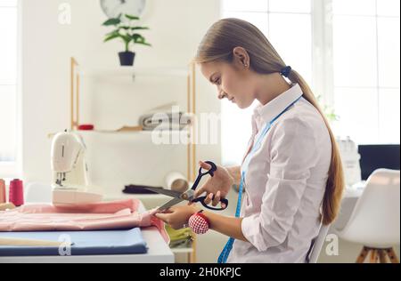 Il seartstress femminile concentrato giovane usa le forbici del sarto per tagliare il tessuto per cucire i vestiti. Foto Stock