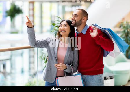 Shopping insieme. Coppia interracial millennial che compra le merci al supermercato, tenendo i sacchetti del compratore, indicando da parte Foto Stock
