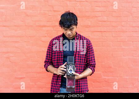 Happy hipster uomo asiatico utilizzando la vecchia macchina fotografica vintage in piedi sul muro rosso - fotografo orientale divertirsi viaggiando per il mondo - gioioso stile di vita giovanile Foto Stock