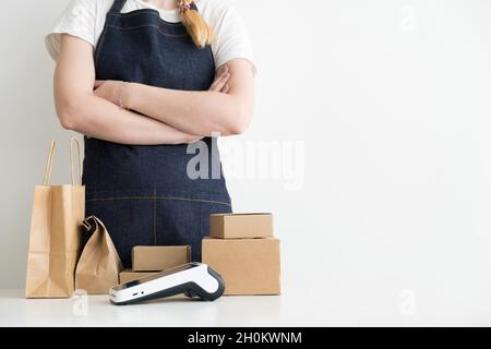 Donna d'affari con le braccia incrociate in piedi dal tavolo con scatole di cartone, confezioni di cartone e moderno terminale POS di pagamento Foto Stock