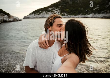 Felice coppia caucasica baciare mentre si prende selfie sulla spiaggia Foto Stock