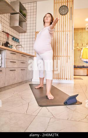 Giovane donna incinta che va yoga esercizio in cucina. Lezioni online di assistenza sanitaria Foto Stock