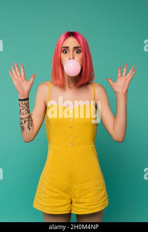 scioccata giovane donna con capelli rosa tinti soffiando bolla gomma isolato su blu Foto Stock