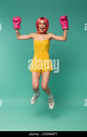 tutta la lunghezza della giovane donna stupita con i capelli tinti rosa nei guanti di boxing che saltano su blu Foto Stock