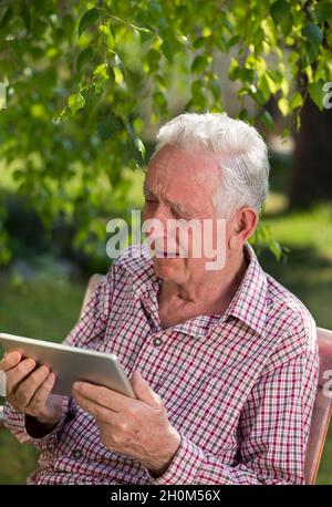 Uomo anziano triste seduto su panca in giardino, guardando il tablet e piangendo Foto Stock