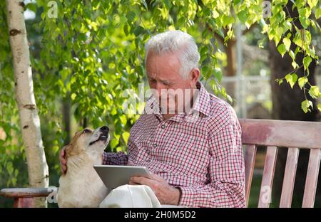 Triste uomo anziano con il suo cane seduto su panca in giardino, guardando il tablet e piangendo Foto Stock