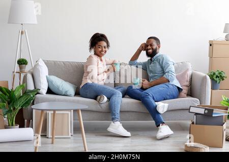 Sorridente uomo e donna afroamericana si rilassa sul divano, bevendo caffè in camera con scatole di cartone Foto Stock