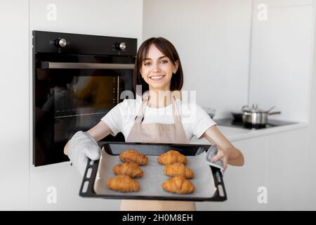 Felice giovane casalinga che tiene vassoio con cornetti appena sfornati, in piedi in cucina e sorridendo alla macchina fotografica Foto Stock