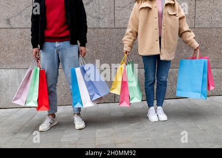 Giovane coppia irriconoscibile che tiene i sacchetti del compratore, posando davanti al muro di pietra all'aperto, andando shopping insieme Foto Stock