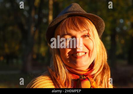 Primo piano ritratto di sorridente giovane bionda caucasica nel colorato parco autunnale. Donna brillante ed elegante in pullover giallo, cappello nero e scialle del collo Foto Stock