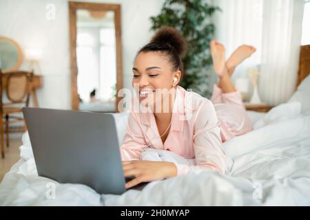 Lavoro in camera da letto. Felice giovane donna afroamericana che lavora su un computer portatile, sdraiato sul letto in una camera da letto accogliente Foto Stock