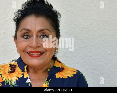 Anziano ottimista positivo fiducioso messicano caucasico Latina donna con il trucco sorride e guarda lo spettatore di fronte a sfondo grigio. Foto Stock