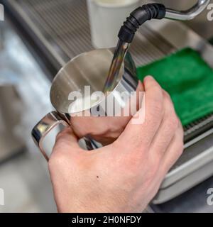 Primo piano delle mani di un barista che prepara il latte caldo su una macchina per il caffè per preparare cappuccino o latte in un bar. Foto quadrata Foto Stock