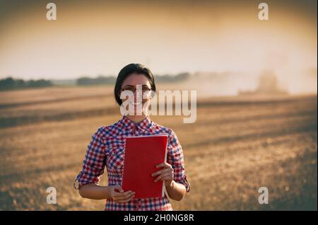 Bella giovane donna con libro di nota in piedi sul campo di grano durante il raccolto. Mietitrebbia che lavora in background Foto Stock