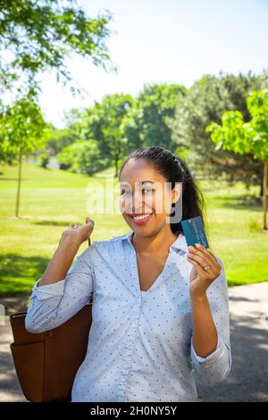 Una donna afroamericana sorridente in una camicia blu con colletto tiene una carta di credito blu in plastica con micro chip con la sua borsetta marrone slung ov Foto Stock