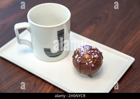 Primo piano: Foto del cibo di una bomba di cacao caldo ricoperta di cioccolato bruno sgocciolato e spolverini viola e bianchi posti su un piatto da portata bianco accanto a A. Foto Stock