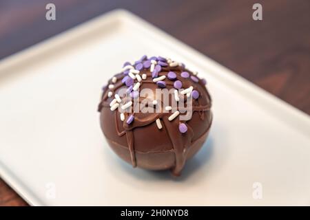 Primo piano: Foto del cibo di una bomba di cioccolata calda o cioccolato ricoperta di cioccolato bruno sgocciolato e spolverini viola e bianchi adagiato su un piano rettangolare Foto Stock