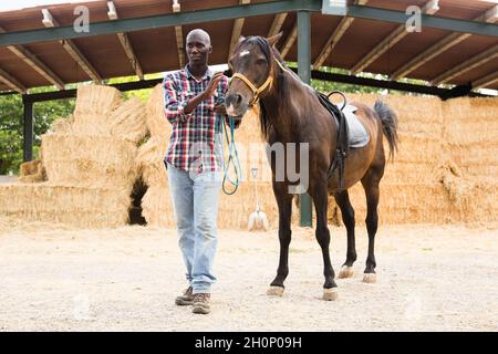 Giovane agricoltore in piedi con cavallo in stalla in fattoria Foto Stock
