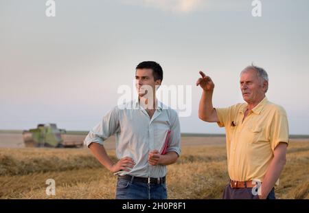 Due agricoltori in piedi sul campo del grano durante la mietitura con la mietitrebbia che lavora in background Foto Stock