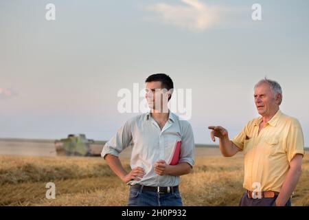 Due agricoltori in piedi sul campo del grano durante la mietitura con la mietitrebbia che lavora in background Foto Stock