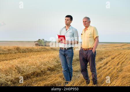 Due agricoltori in piedi sul campo del grano durante la mietitura con la mietitrebbia che lavora in background Foto Stock