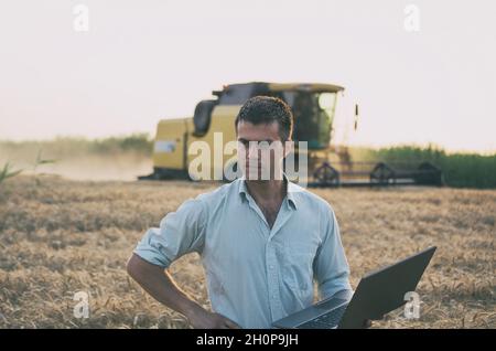 Giovane ingegnere contadino con computer portatile in posizione verticale sul campo del grano mentre la mietitrebbia lavora in background Foto Stock