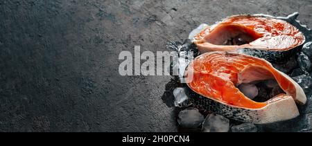 Due bistecche di salmone selvatico crudo su ghiaccio su sfondo nero testurizzato. Bistecche di salmone con spazio di copia a sinistra. Banner orizzontale lungo Foto Stock