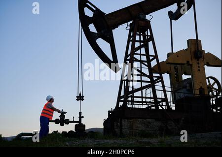 Operatore petrolifero in gilet da lavoro che utilizza l'unità di pompaggio per estrarre il petrolio grezzo dal pozzo petrolifero. L'oleatore aziona il martinetto della pompa del petrolio mentre lavora nel campo dell'olio la sera. Concetto di estrazione dell'olio. Foto Stock