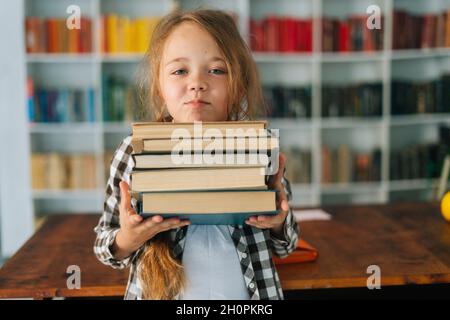 Ritratto medio del colpo della ragazza elementare graziosa della scuola del bambino che tiene la pila dei libri nella biblioteca alla scuola che guarda la macchina fotografica. Foto Stock