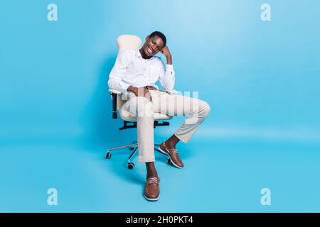 Foto del ragazzo piuttosto premuroso pelle scura vestito formale camicia seduta sedia sorridente isolato colore sfondo blu Foto Stock