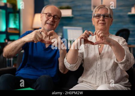 Sposata vecchia coppia che fa il simbolo del cuore con le mani mentre guarda la macchina fotografica nel soggiorno. Marito e moglie in pensione creano un romantico gesto d'amore seduto sul divano di casa. Rapporti senior Foto Stock